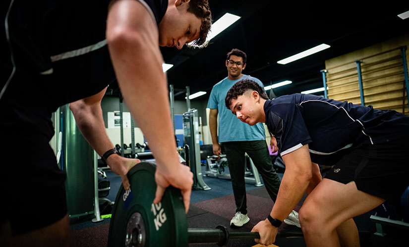 Two sport and exercise students load up weights on a barbell for a male client waiting to do a deadlift.