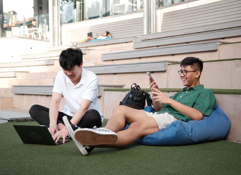 Two male students sit together, one is looking at a laptop the other is looking at his phone.