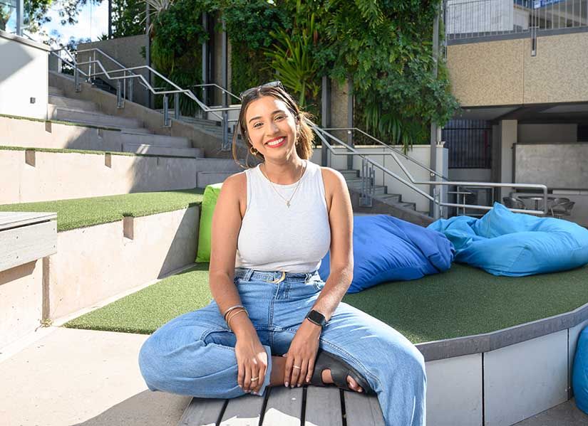 Smiling female student in jeans and a casual top sits on campus in the sunshine.