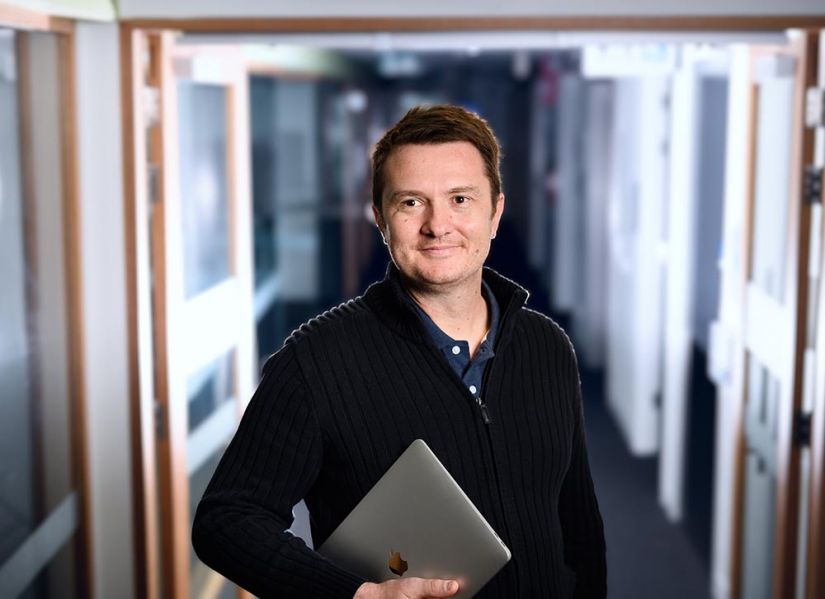 A man in a dark, collared sweater stands in an office hallway. He's holding a laptop and smiling at someone off camera.