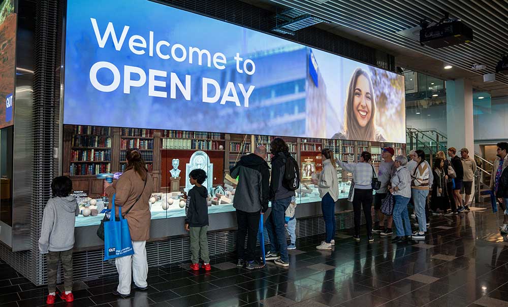 A crowd of people stands in front of a large, interactive display screen. At the top of the screen is a digital banner displaying the words 'Welcome to Open Day'.