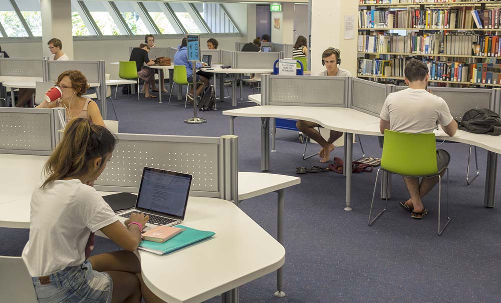 A quiet study area on campus. Students sit at desks with books and lap tops. One student takes a sip from a takeaway coffee cup.