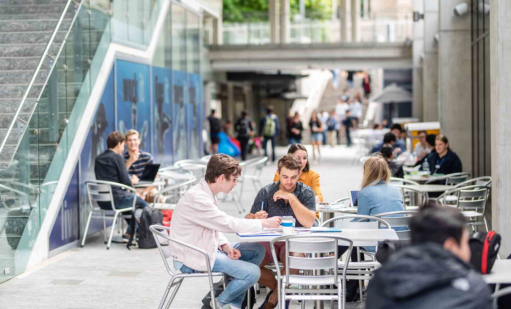 A busy campus foodcourt with groups of students sitting at tables chatting, studying and relaxing while they eat.
