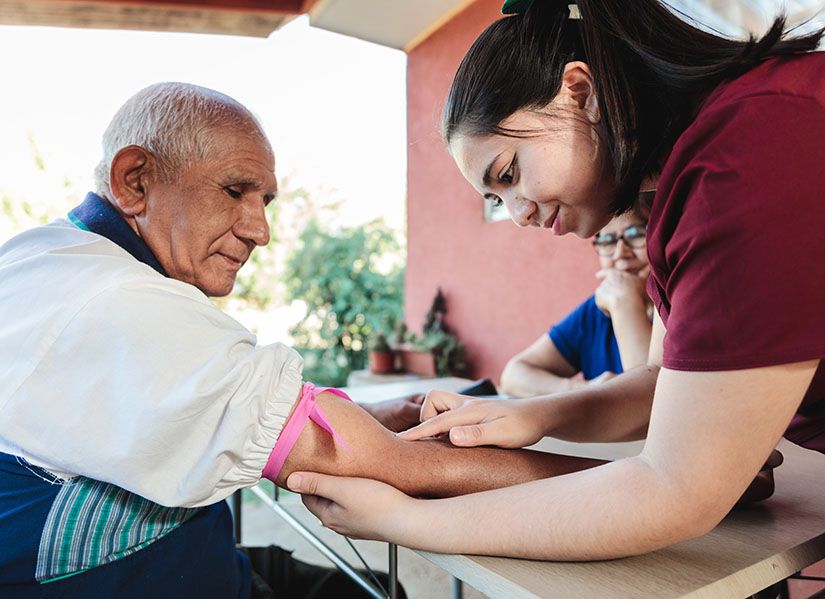 A female nurse in scrubs checks a male patient's arm for a vein during a home visit.