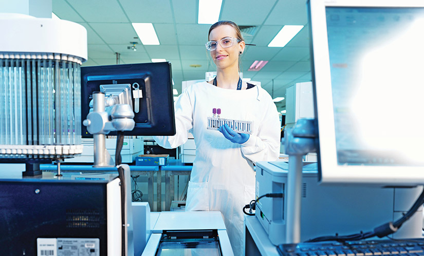 A woman in a labcoat holding a tray of samples stands at a lab bench.