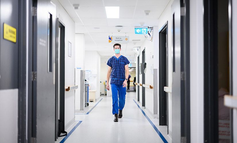 A man in scrubs walks with purpose through a hospital hallway.