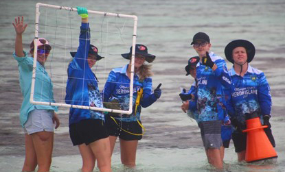 A smiling group of students and teachers standing knee deep in the ocean and shouldering equipment wave at the camera.