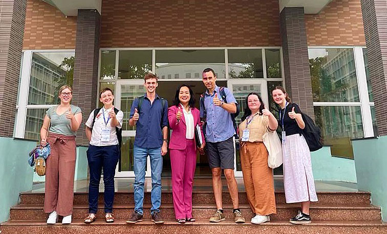 Two professors and a group of students stand on the steps of a university building giving the thumbs up.