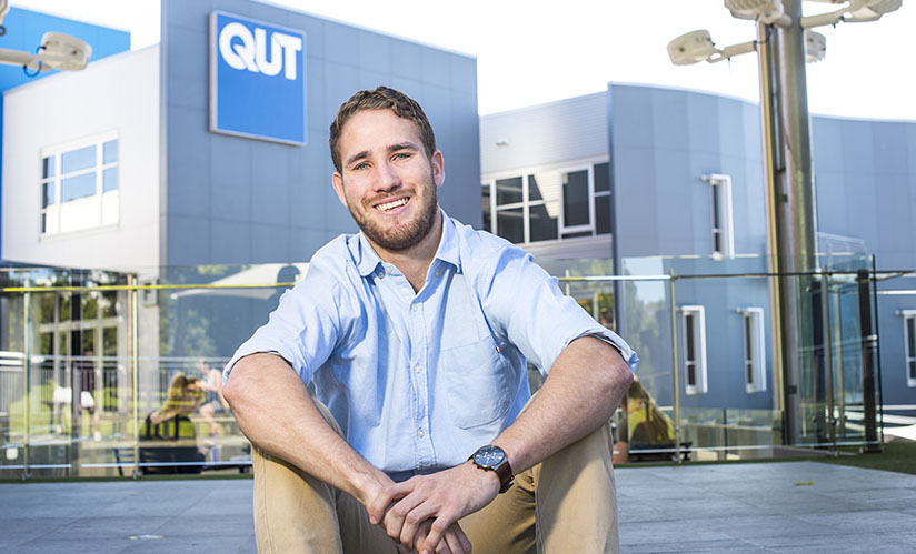 A smiling man wearing a business attire stands in front of a modern, office-like building on a leafy campus.
