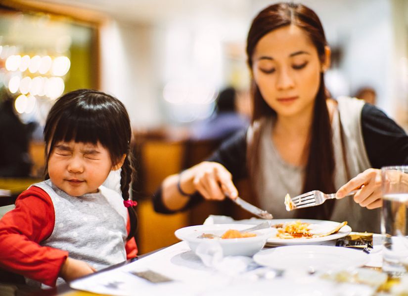 A toddler squints her eyes shut and turns away while her mum cuts up her food.