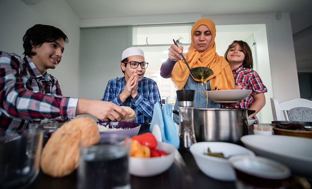 A mother in a bright, orange headscarf ladles soup from a pot into a bowl and three excited, hungry boys break pieces of bread while they wait.