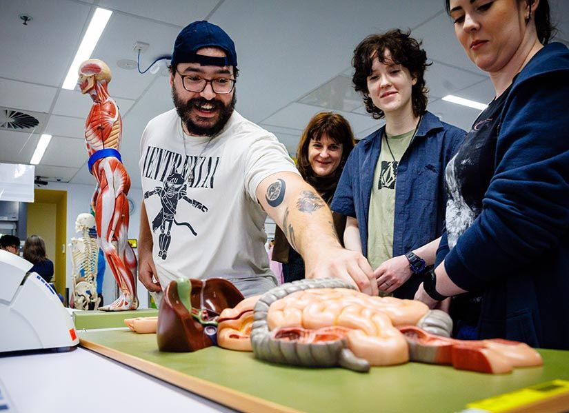 A bearded man smiles as he points out the features of an anatomical model to two interested students.
