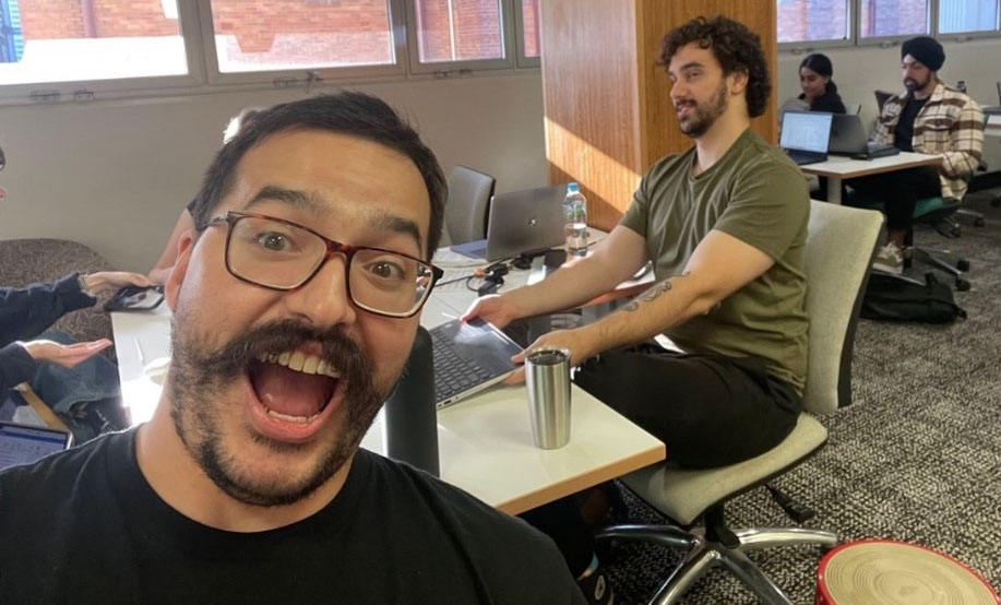 Two students sit at a desk that is covered in text books, note pads, a laptop and water bottles. One is laughing while the other is grimacing.