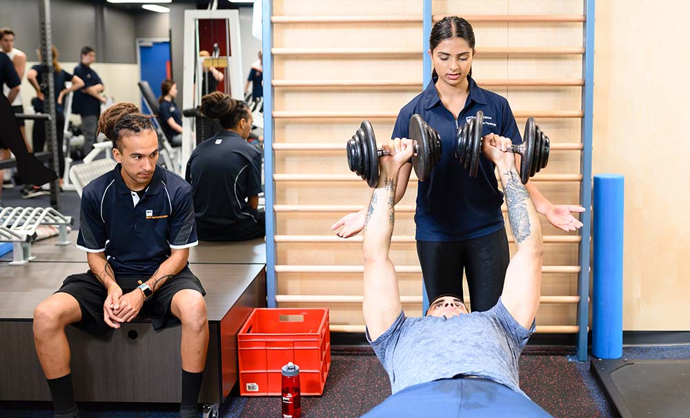 A male client lies on the bench lifting free weights while two exercise science students look on.