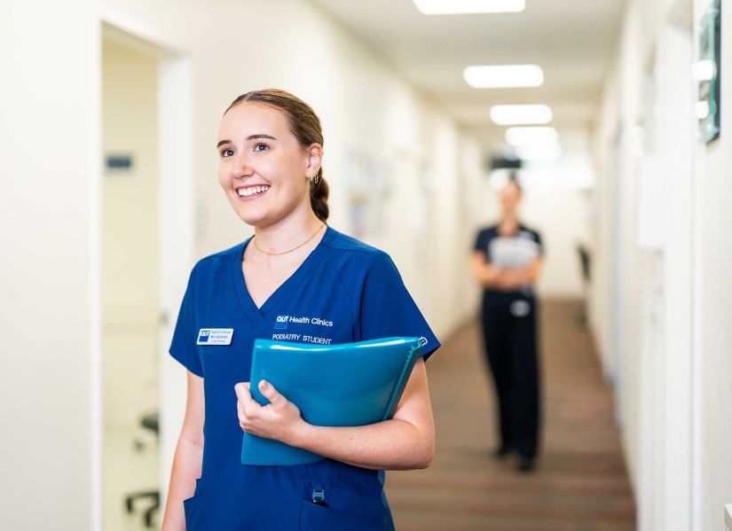 A female student in scrubs holding a patient file walks down the hallway of a clinic towards a consultation room.