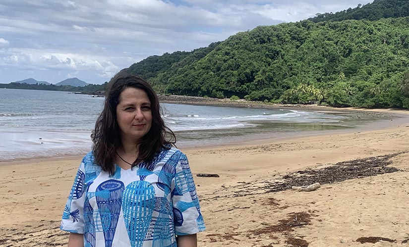 A woman walks along an empty beach. Behind her mountains covered in tropical rainforest rise behind the sands of the beach.
