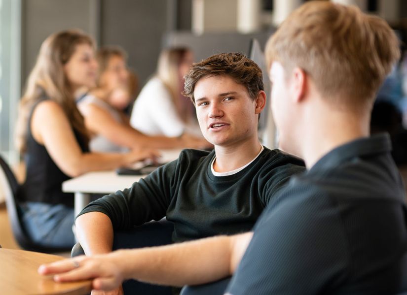 A male student sits at a table with another student. He is measuring his words as he responds to something that has just been said.