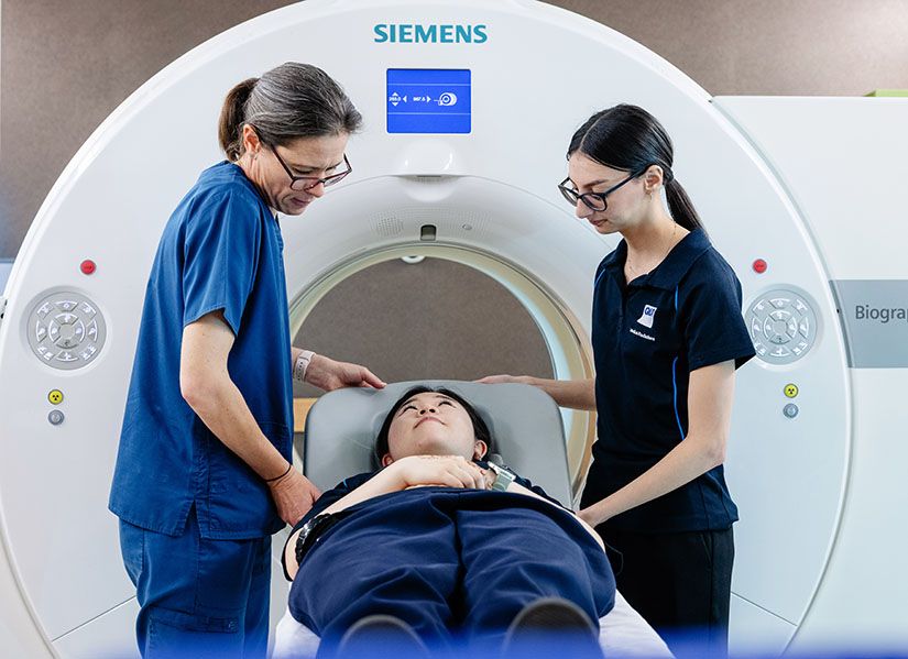 A student and a techician stand either side of an MRI machine. Another student lies on the patient table as part of a training exercise.