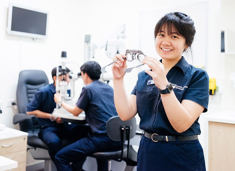 A female optometry student holds glasses for eye tests while two other students practice giving an eye exam.