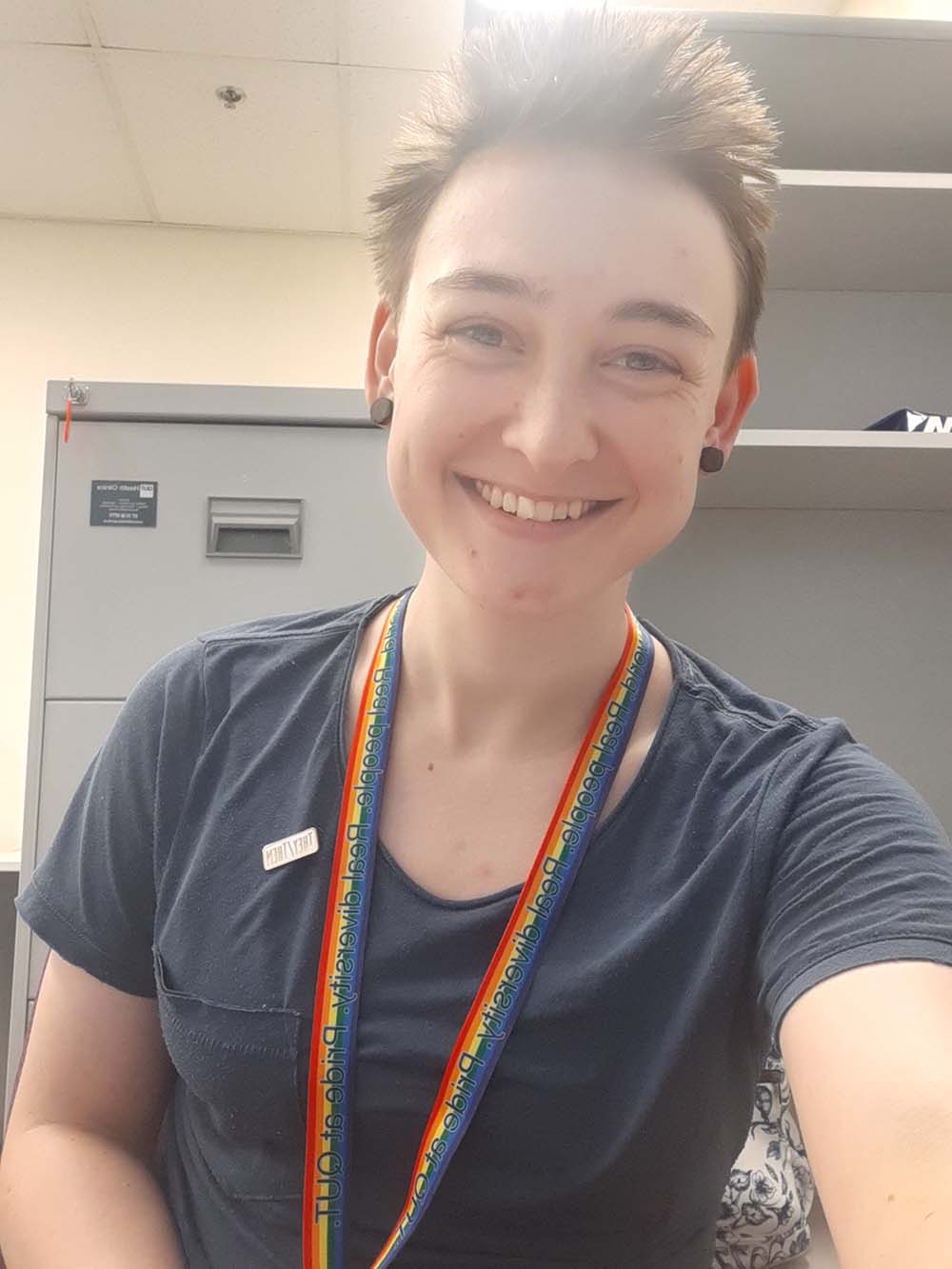 A smiling student wearing a rainbow lanyard sits in a research office.