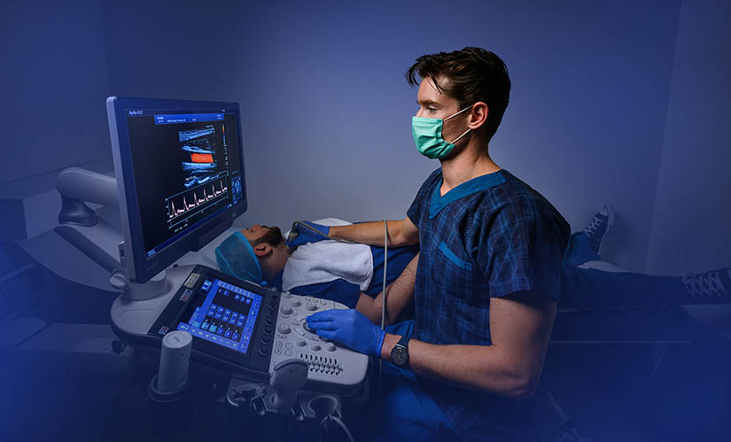 A man in scrubs sits in front of a computer screen while holding equipment up to a patient lying in a hospital bed.