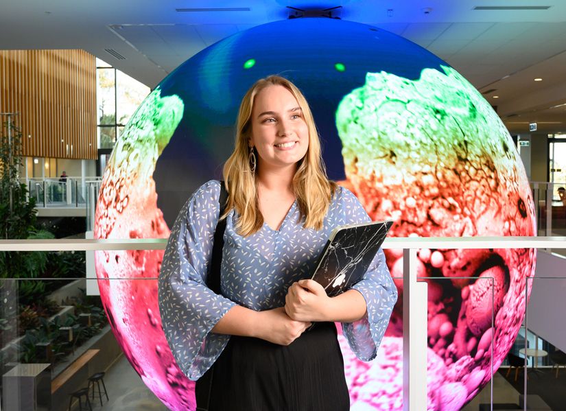 Female student Nicola holding her laptop standing in front of a large digital sphere projecting a coral reef