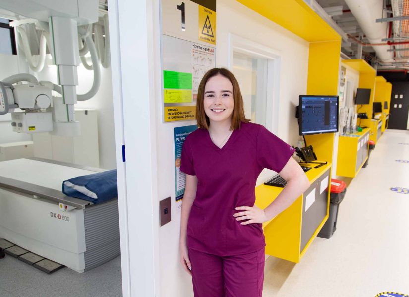 A woman in maroon scrubs stands in the doorway of a room filled with medical imaging equipment and a hospital bed.