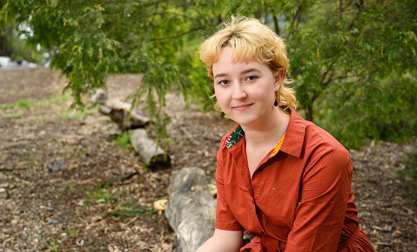 A young woman with a blonde mullet and collared shirt sits outdoors in a country setting.