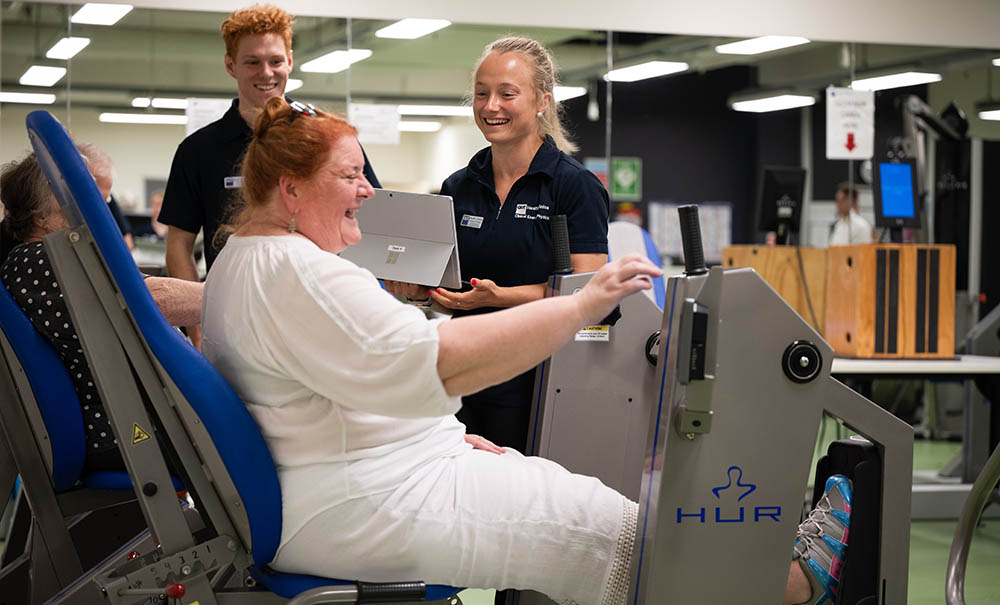 An older woman using a leg press machine laughs as she shares a joke with two exercise physiology students supervising her.