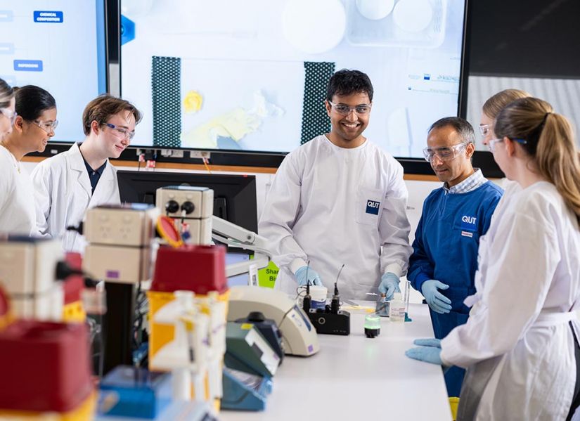 A male student in a labcoat and protective glasses stands at a lab bench. In front of him is cream that he is mixing, standing next to him is a tutor and a group of fellow students.