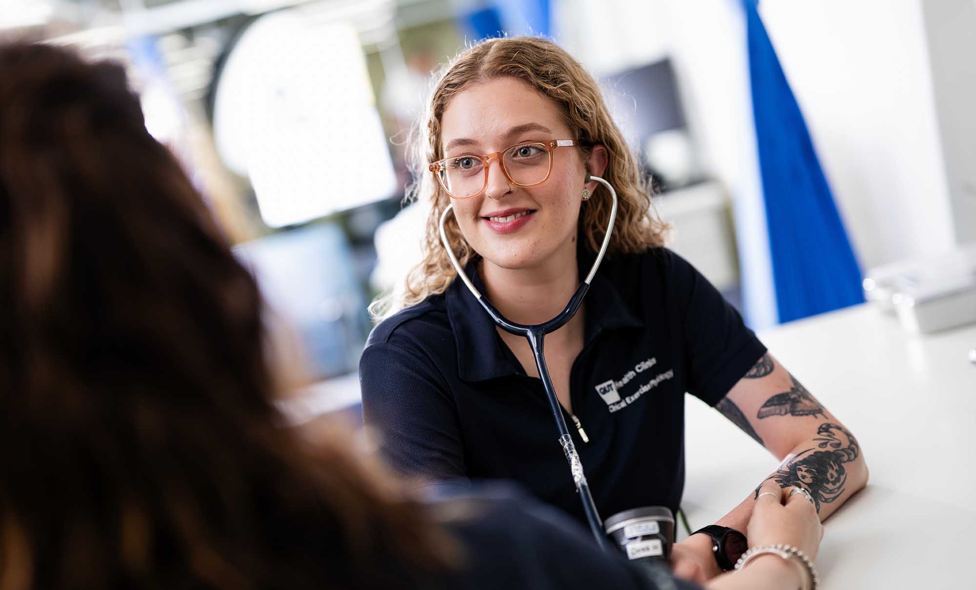 A female students wearing a uniform, with traditional tattoos on her arm, listens to a stethoscope as she takes a client's blood pressure.