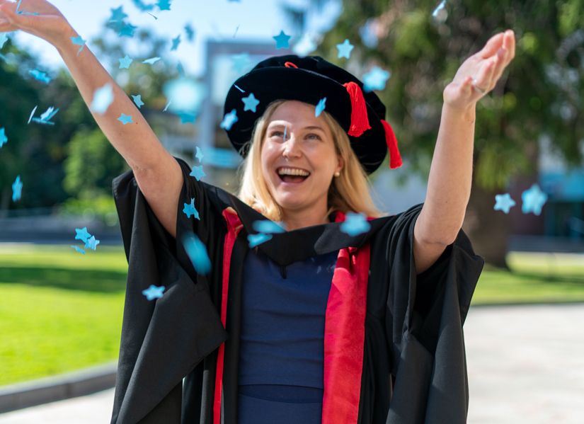 Happy postgraduate student wearing her graduation gown, throwing blue confetti in the air