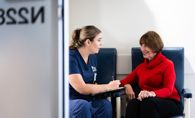 A nurse in hospital scrubs takes notes on a clipboard while talking with a female patient.
