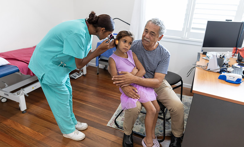 A female nurse performs a check in the ear of a little girl while she sits on her father's lap. Both patient and nurse are Aboriginal and are relaxed and smiling.