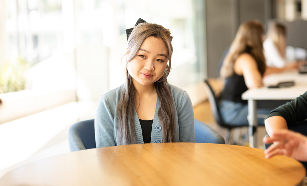 A female, Asian wearing a large bow in her hair sits at a wooden desk with other students. She is listening intently to what is being said.