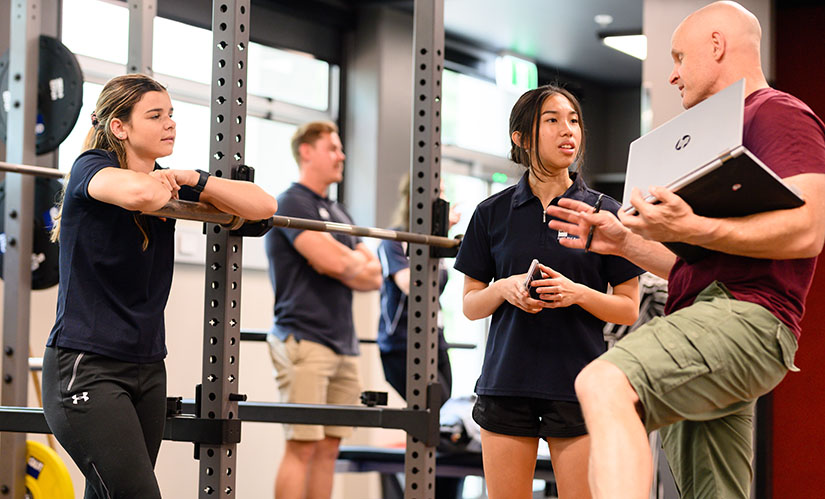 Two female students in sports uniforms lean on equipment in a gym while listening to a lecturer explain a concept.