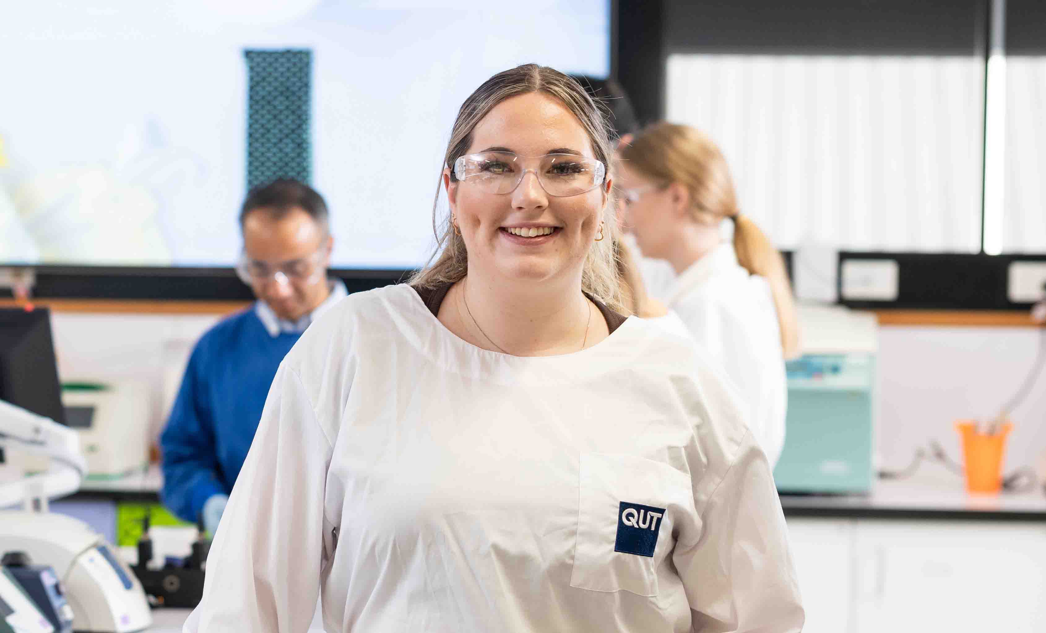 A smiling, blonde, female pharmacy student wearing a lab coat and glasses stands in a laboratory.