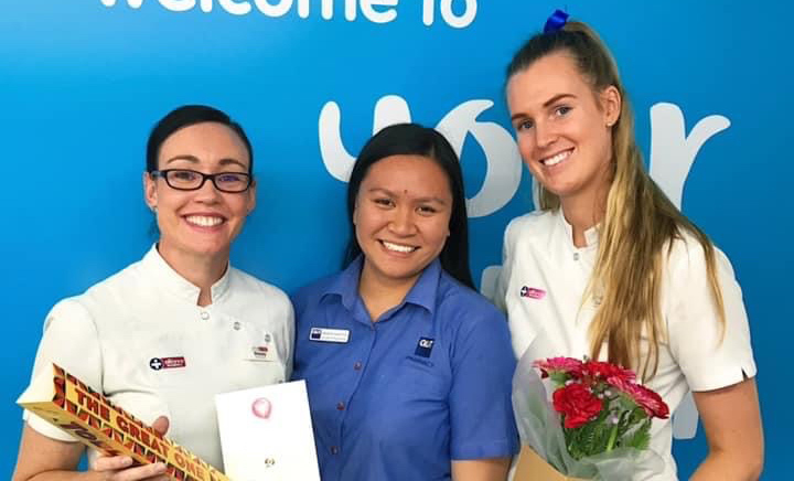Three smiling women standing in clinical uniforms at a pharmacy.