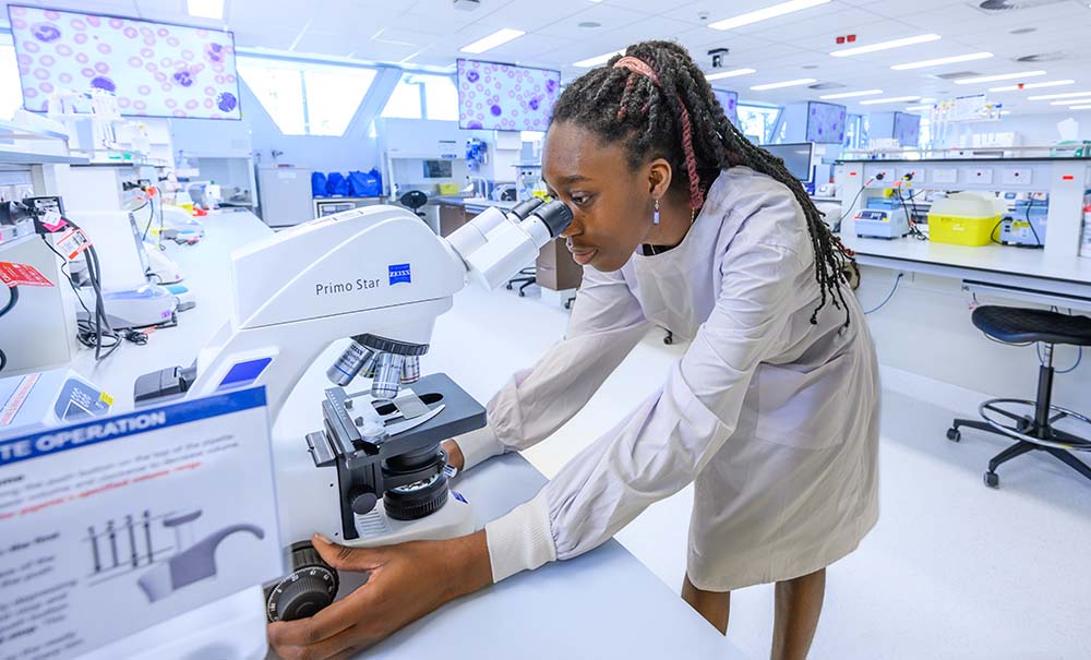 Abena, wears a lab coat and has her braids tied up in a pony tail. She adjusts the dial on a microscope as she peers down the lens at a slide.