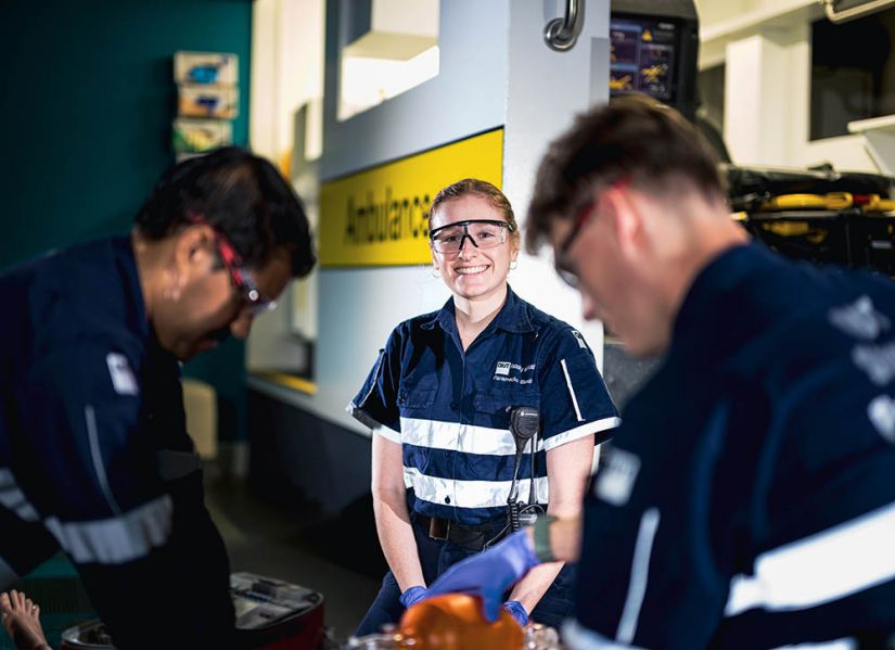 Three students in paramedic uniforms practice reviving a dummy in class. Megan, a female, red-headed student pauses and looks up to smile at the camera.
