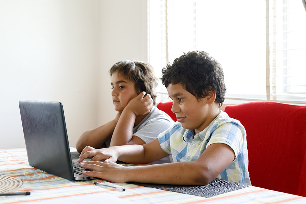 Two young boys sit at a kitchen table looking up information on a lap-top.