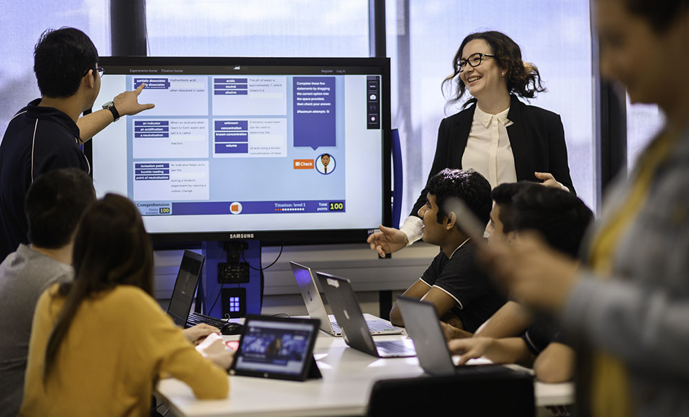 A female guest lecturer in glasses and a jacket smiles as she watches a student demonstrate an IT process on a large screen. Students with laptops sit at a table and watch the lesson.