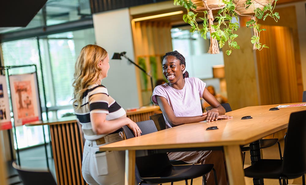Abena sits at a large wooden table in an open learning space. She smiles at a friend who is joining her and is about to take her seat.