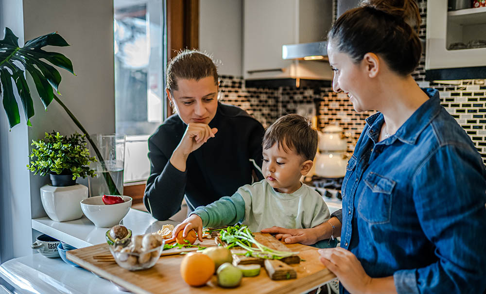 Two mums stand at the kitchen bench. Their toddler stands on a booster chair between them, he's helping to sort vegetables for chopping.