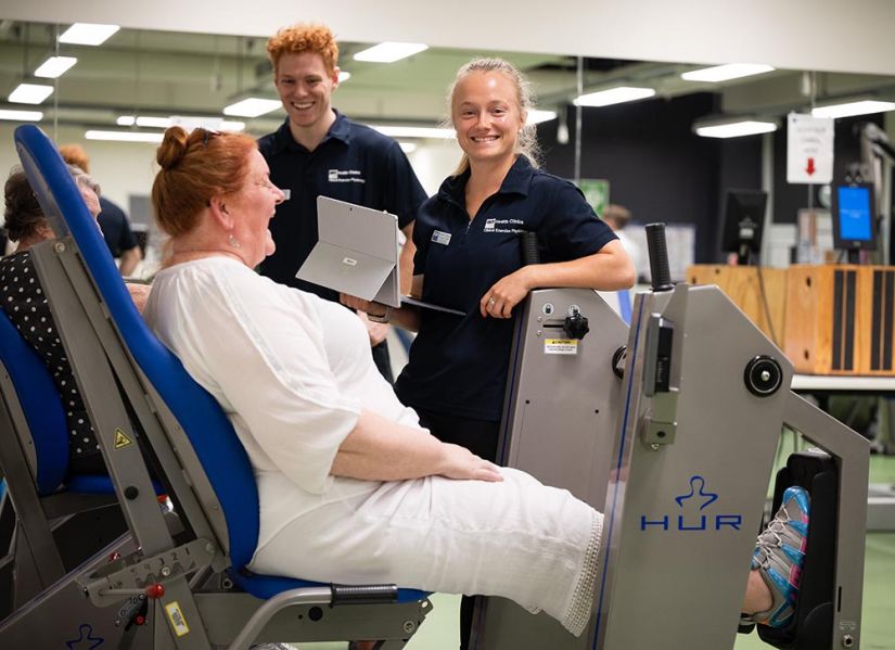 Two smiling students stand behind exercise equipment while a client using the leg press machine infront of them has a laugh.