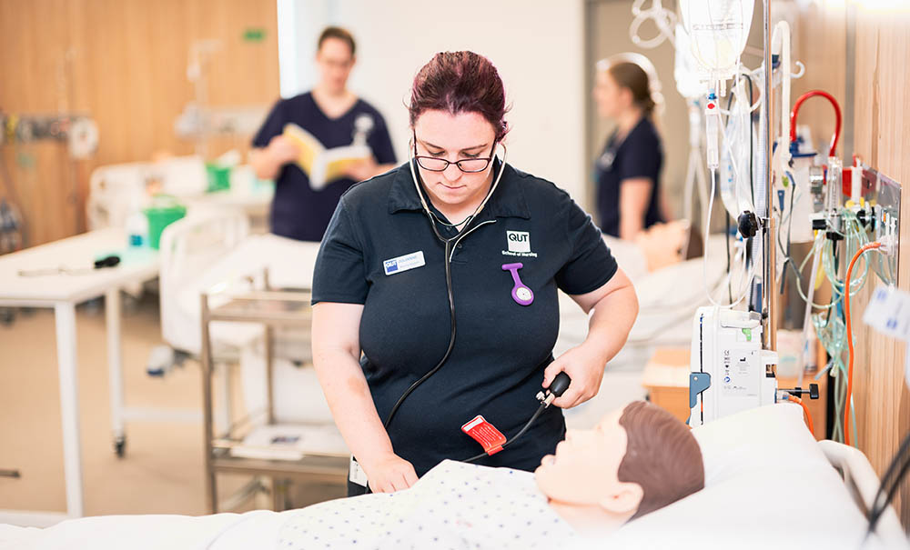 A female student in a nursing uniform looks down at the equipment on the table in front of her. She is standing in a simulated hospital ward and is about to run a scenario on the dummy lying in the hospital bed next to her.