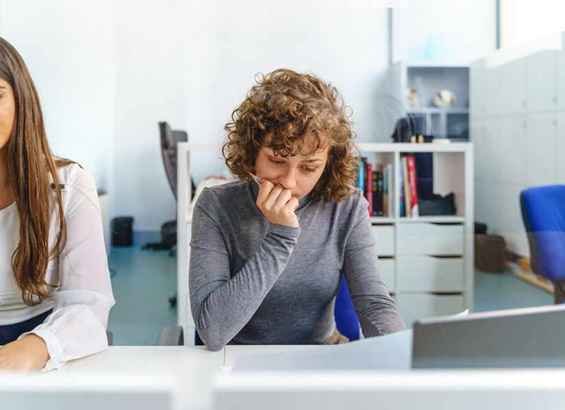 A young woman sits at a desk in a clinical setting. She looks worried as she stares at the paper she is holding.