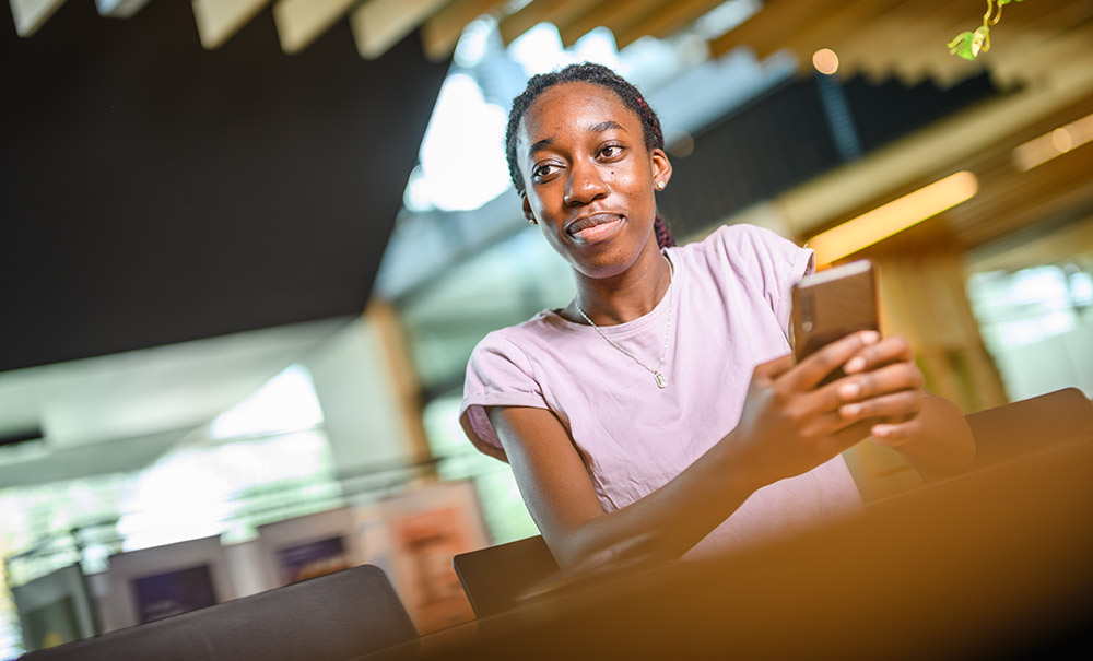 Abena wears a casual shirt. She sits at a table with a phone in her hand, she's staring into the distance with a thoughtful look on her face.