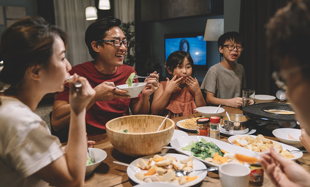 A father sits at a family table surrounded by two kids, a mother and a grandmother. He's holding a bowl of vegetables and a fork close to his face and is laughing at a joke someone has made.