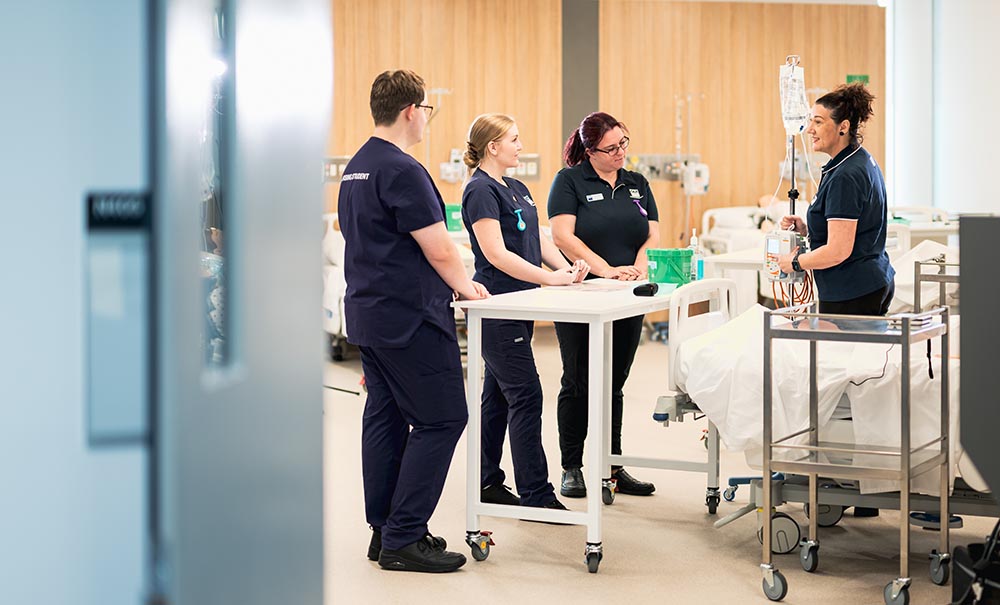 Three nursing students stand in a simulated hospital ward on campus, they are listening to a nurse academic who is explaining a technique.